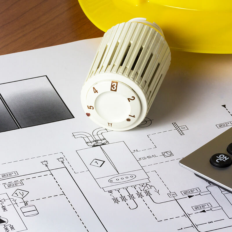 Ein Heizungsthermostat, Pläne und mehr auf einem Schreibtisch – symbolische Darstellung für die Heizlastberechnung und den hydraulischen Abgleich durch die Wüstenrot Energieberatung